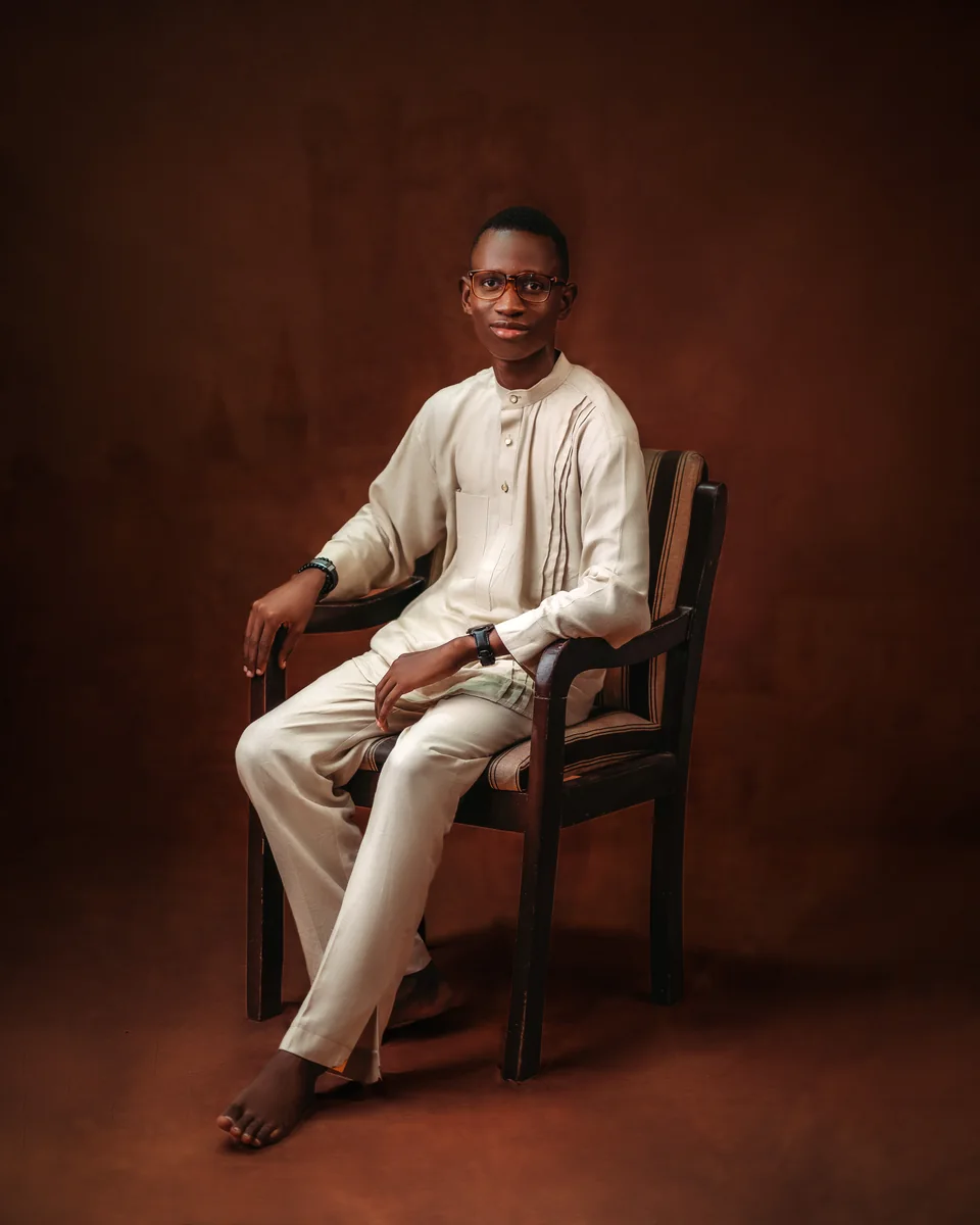 Divine Jemegbe seated in a cream outfit against a warm brown studio backdrop.